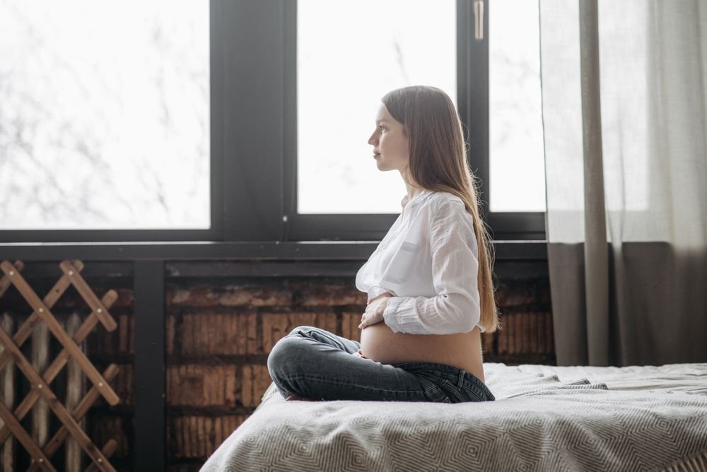Gestante sentada na cama com as mãos sobre a barriga